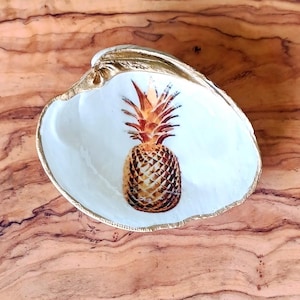 Puede incluir: Una concha de mar blanca con un borde dorado, que presenta una imagen pintada de una piña con tonos marrones y amarillos.