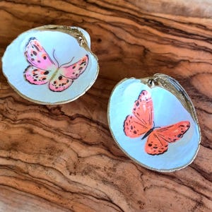 May include: Two seashells with painted butterflies. The seashells are white with a gold trim. One butterfly is pink with black spots, the other is orange with black spots.