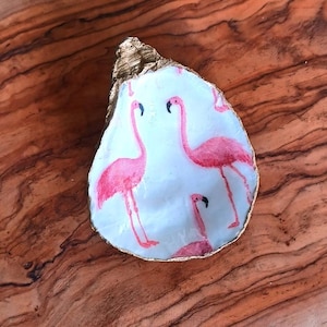 May include: A white oyster shell with a gold rim, painted with pink flamingos. The shell is a decorative trinket or dish.