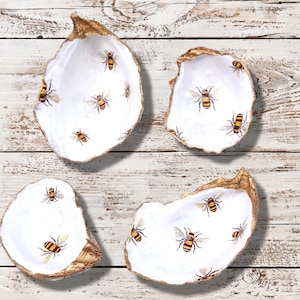 May include: Four white oyster shell dishes with a gold rim, each decorated with multiple illustrations of bees. The bees are yellow and black. The shells are arranged on a white wooden surface.