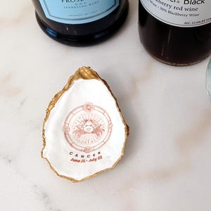May include: A white oyster shell with gold trim, featuring a pink and white illustration of the Cancer zodiac sign with the text "CANCER June 22 - July 22".