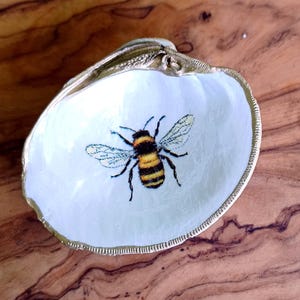 May include: A white seashell with a gold rim, painted with a yellow and black bumblebee.