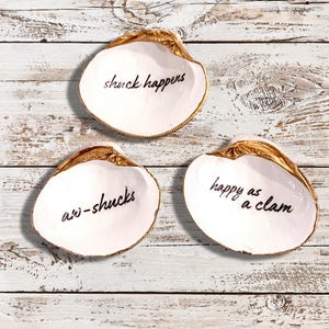 Pode incluir: Três pratos de cerâmica branca em forma de concha de amêijoa com acabamento dourado. Os pratos têm as seguintes frases escritas neles: "shuck happens", "aw-shucks" e "happy as a clam".