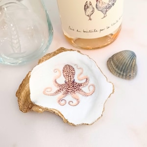 May include: A white oyster shell dish with a gold rim, featuring a watercolor octopus design. A bottle of wine with a chicken illustration and a seashell are also present. A clear glass is in the background.
