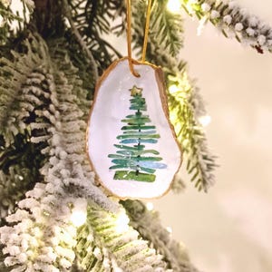 May include: A Christmas tree ornament made from a natural oyster shell. The shell is painted white inside with a green and blue Christmas tree design and a gold star. A gold string is attached for hanging. The ornament hangs on a snowy Christmas tree.