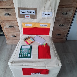 May include: A beige fabric play chair with a red, yellow, and beige post office theme. The chair has pockets labeled "Money", "Stamps", and a red pocket for letters. It also has a calculator, notepad, and pencils.