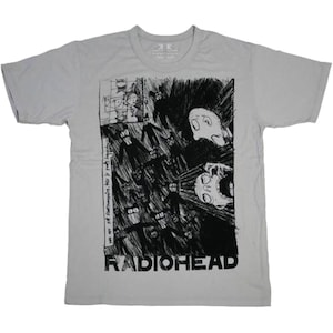 Könnte beinhalten: Hellgraues T-Shirt mit einem schwarz-weißen Grafikdesign. Das Design beinhaltet eine Illustration im Comic-Stil und den Bandnamen "RADIOHEAD" in fetten Buchstaben. Das T-Shirt hat einen Rundhalsausschnitt und kurze Ärmel.