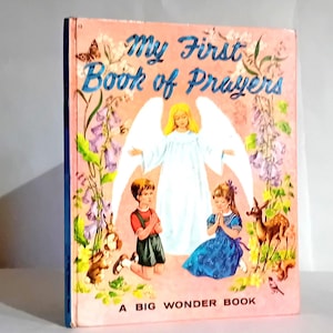 Puede incluir: Un libro rosa con el título "My First Book of Prayers" y una ilustración de un ángel con dos niños rezando. El libro es un "Big Wonder Book".