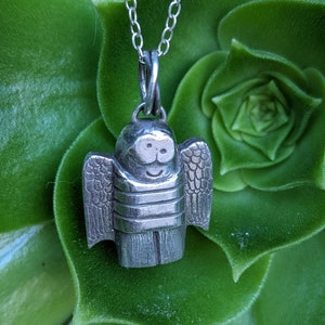 a dainty necklace of a hand carved sterling silver angel child with wings, cartoon eyes and a sweet smile dressed in a striped shirt and pants strung on a cable chain hanging in front of a large green succulent