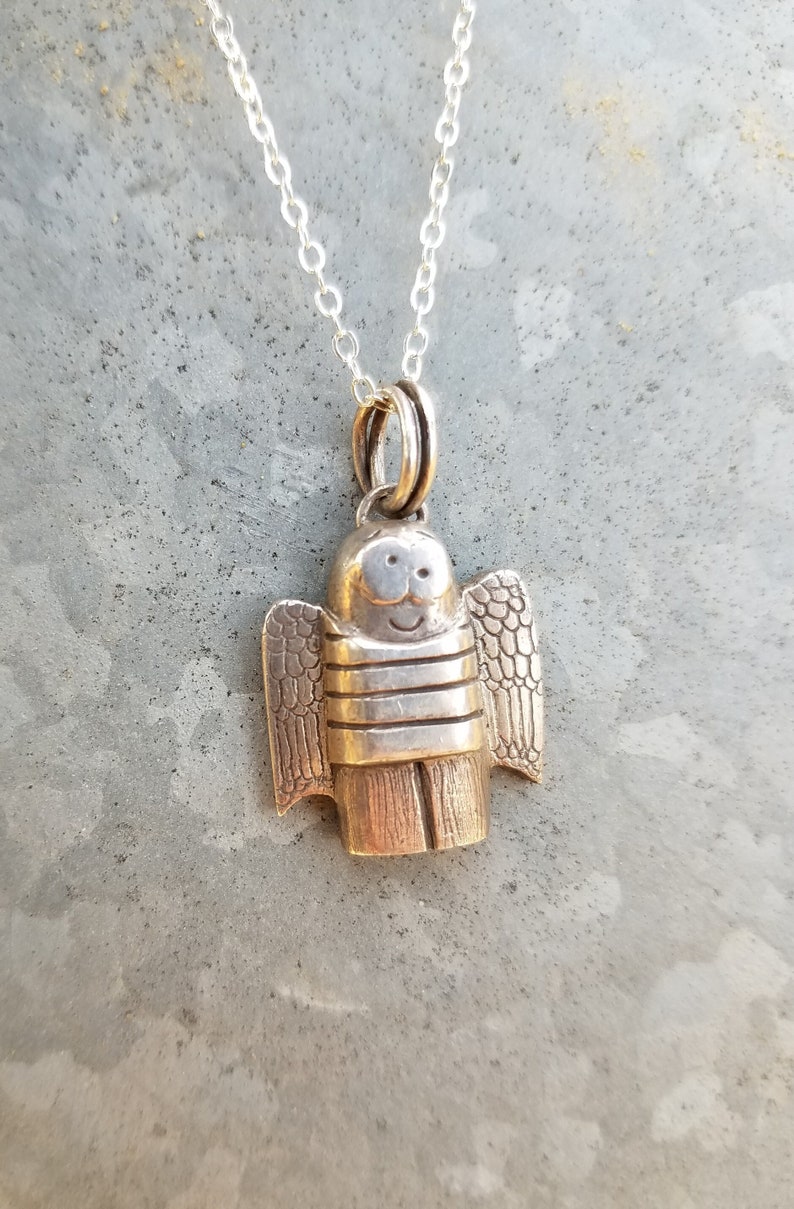 a dainty necklace of a hand carved sterling silver angel child with wings, cartoon eyes and a sweet smile dressed in a striped shirt and pants strung on a cable chain draped over patterned metal