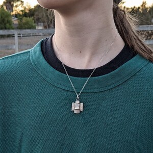 a dainty necklace of a hand carved sterling silver angel child with wings, cartoon eyes and a sweet smile dressed in a striped shirt and pants shown on a model in green and black shirt in a farm setting