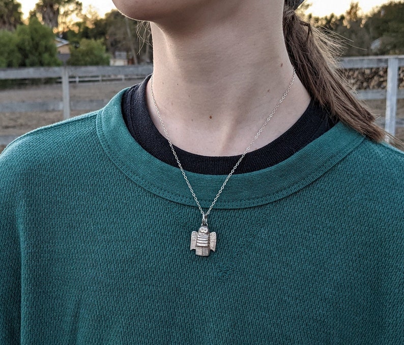 a dainty necklace of a hand carved sterling silver angel child with wings, cartoon eyes and a sweet smile dressed in a striped shirt and pants shown on a model in green and black shirt in a farm setting