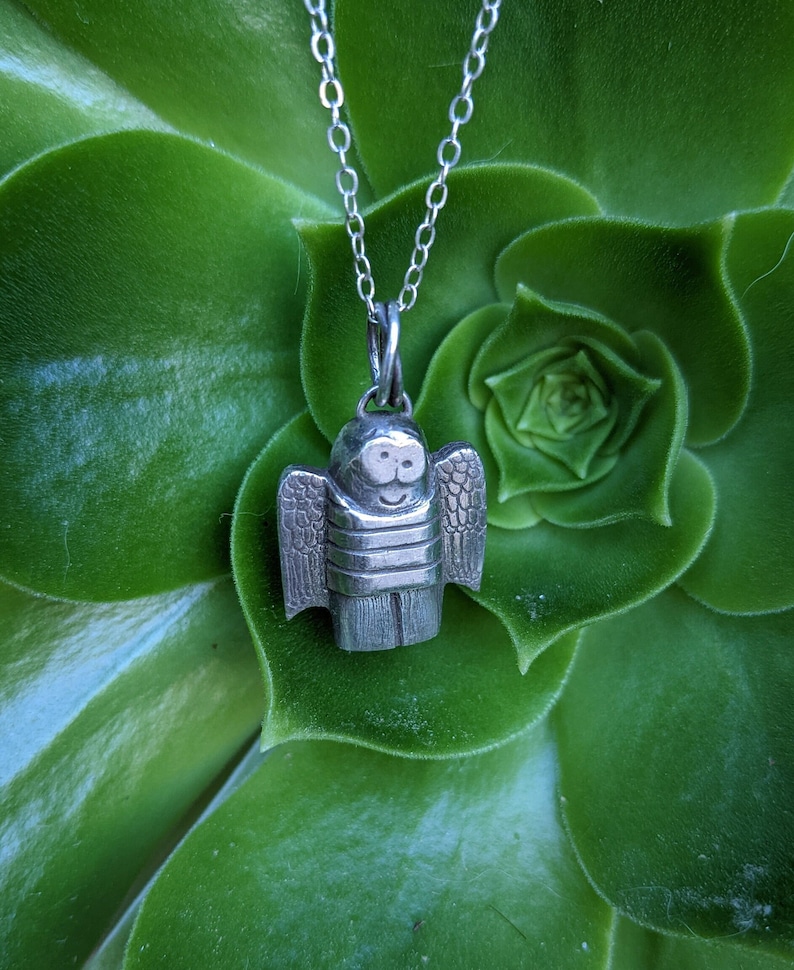 a dainty necklace of a hand carved sterling silver angel child with wings, cartoon eyes and a sweet smile dressed in a striped shirt and pants strung on a cable chain hanging in front of a large green succulent