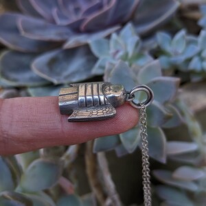 the side view of a dainty necklace of a hand carved sterling silver angel child with wings, cartoon eyes and a sweet smile dressed in a striped shirt and pants strung on a cable chain draped over a finger