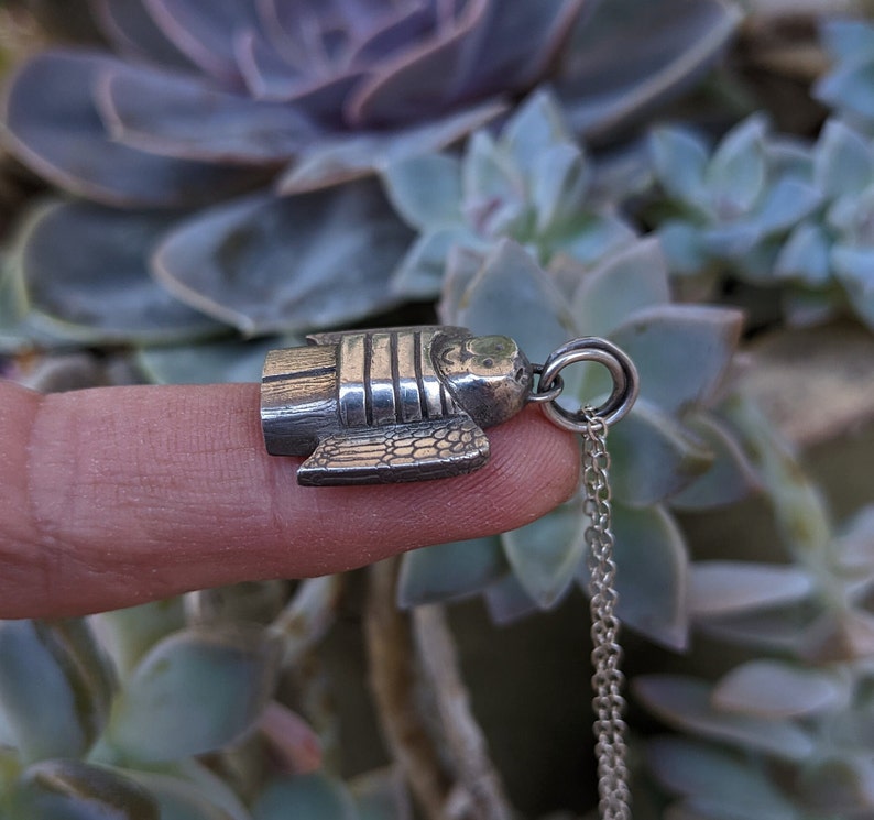 the side view of a dainty necklace of a hand carved sterling silver angel child with wings, cartoon eyes and a sweet smile dressed in a striped shirt and pants strung on a cable chain draped over a finger