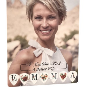 May include: A personalized gift featuring a smiling woman with blonde hair, wearing a white top with a bow. Below the image are guitar picks spelling "EMMA" and "A", with floral and rose designs. The text reads "I Couldn't Pick A Better Wife".