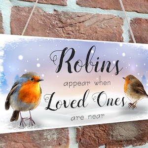 Op de afbeelding: Een wit houten bord met een winterse achtergrond waarop twee roodborstjes te zien zijn. Het bord luidt "Roodborstjes verschijnen wanneer geliefden in de buurt zijn".