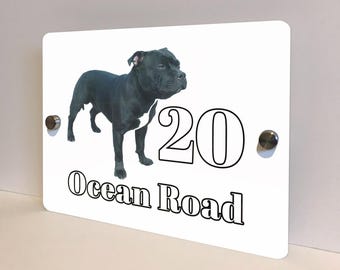 May include: A white rectangular house sign with a black dog illustration and the text "20 Ocean Road". The dog is black with a white chest. The numbers and text are in black. The sign is mounted with two silver screws.