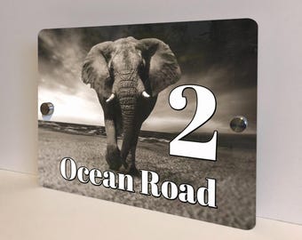May include: A rectangular house number sign featuring a sepia-toned image of an elephant walking towards the viewer. The number "2" and the words "Ocean Road" are in white, bold font. The sign has two silver mounting studs.