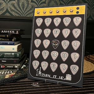 May include: A black rectangular advent calendar featuring a yellow amplifier control panel at the top with knobs and settings. The board displays twenty-four guitar pick-shaped slots numbered one through twenty-four, with the text TURN IT UP.