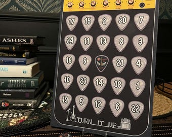 May include: A black rectangular advent calendar featuring a yellow amplifier control panel at the top with knobs and settings. The board displays twenty-four guitar pick-shaped slots numbered one through twenty-four, with the text TURN IT UP.