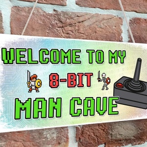 May include: A white wooden sign with a pixelated design. The sign says "Welcome to my 8-bit Man Cave" with a pixelated image of a knight and a joystick.