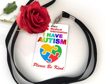 Puede incluir: Una tarjeta blanca con el texto "Información médica TENGO AUTISMO Por favor, sé amable" y un corazón de pieza de rompecabezas arcoíris. La tarjeta está unida a un cordón negro con una rosa roja.