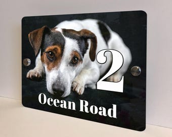 May include: A rectangular black house number sign featuring a Jack Russell Terrier dog. The number "2" is in white, along with the words "Ocean Road". The sign is mounted with silver hardware.