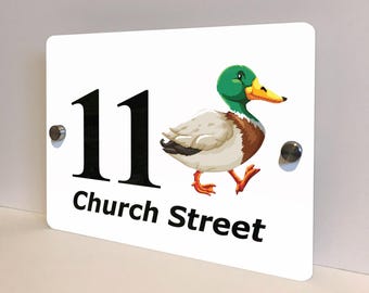 May include: White rectangular house number sign with the number 11 in black, and the words "Church Street" in black. A colorful illustration of a duck is on the right side of the sign. The sign is mounted with two silver screws.