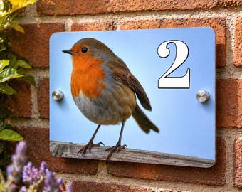 May include: A rectangular house number sign featuring a detailed illustration of a European robin with a bright orange breast, perched on a wooden branch. The number "2" is displayed in white. The sign is mounted on a brick wall.