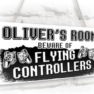 May include: A white sign with black text and graphics that reads "Oliver's Room Beware of Flying Controllers". The sign has a distressed look and features two black and white video game controllers.