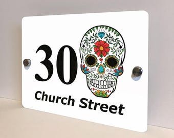 May include: White rectangular house number sign with the number "30" in black and the words "Church Street" below. A colorful sugar skull design is on the right side of the sign. The sign is mounted with two silver screws.