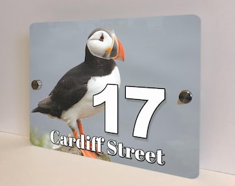 May include: Rectangular address sign featuring a colorful puffin bird illustration. The sign displays the number "17" in large white font, along with the street name "Cardiff Street" in a stylized font. The background is a light gray color.