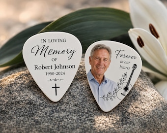 May include: Two white guitar picks featuring memorial designs. One pick displays the text IN LOVING MEMORY OF ROBERT JOHNSON 1950-2024 with a cross. The other pick features a portrait of a man, a guitar illustration, and the text Forever in our hearts.