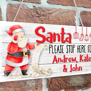 Op de afbeelding: Een wit houten bord met een cartoon afbeelding van de kerstman die zijn lijst controleert. Het bord staat er "Santa PLEASE STOP HERE FOR Andrew, Kate & John".