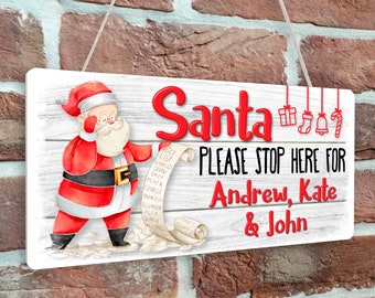 May include: A white wooden sign with a cartoon image of Santa Claus checking his list. The sign reads "Santa PLEASE STOP HERE FOR Andrew, Kate & John".