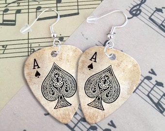 May include: A pair of earrings shaped like guitar picks, featuring the Ace of Spades playing card design. The earrings have a vintage, distressed look with a black spade symbol and the letter "A". They hang from silver-colored ear wires.