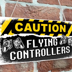 Puede incluir: Un cartel de precaución amarillo y negro con el texto "CAUTION FLYING CONTROLLERS" en blanco. El cartel tiene un aspecto desgastado y presenta un diseño pixelado. Hay imágenes de mandos de videojuegos en el cartel.