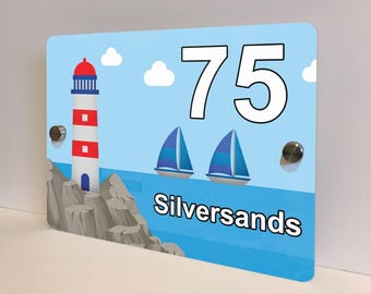 May include: A rectangular sign with a blue background features a lighthouse, sailboats, and the number 75. The word "Silversands" is written across the bottom. The lighthouse is red, white, and blue. Two silver screws are visible.