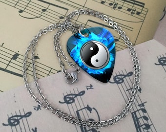 May include: A silver-colored necklace with a guitar pick-shaped pendant. The pendant features a Yin and Yang symbol in black and white, set against a blue floral background. The necklace rests on a sheet of music.