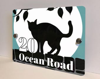May include: A rectangular address sign featuring a black cat silhouette against a white circle, with a teal background. The sign displays the number "20" and the words "Ocean Road" in white. The design includes a branch with leaves.