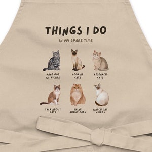 May include: A beige kitchen apron featuring a graphic print of cats and the text "THINGS I DO IN MY SPARE TIME". The graphic depicts six cats with text below each cat describing activities such as "Hang out with cats", "Look at cats", "Research cats", "Talk about cats", "Think about cats", and "Watch cat videos".