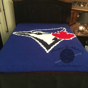 Op de afbeelding: Blauwe gehaakte deken met een wit en zwart Toronto Blue Jays logo. Het logo bevat een rood esdoornblad. De deken heeft een witte rand en de tekst "Megan & Lana's Crochet Creations" staat in de rechterbenedenhoek.
