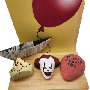 May include: Three bath bombs on a wooden platform. The bath bombs are shaped like a clown face, a red balloon, and a triangle. The balloon bath bomb has the text "You'll Float Too" written on it.