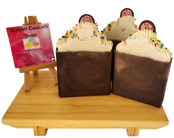 May include: Sugar cookie soaps with white frosting, colorful sprinkles, and a cookie on top. The soaps are dark brown and sit on a wooden tray with a small easel displaying the product name and ingredients.