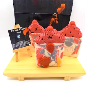 May include: Three handmade soap bars with a red and white swirl design. The soap bars are topped with red frosting and a red balloon with the words "We All Float" on it. The soap bars are on a wooden tray with a small easel holding a sign that says "We All Float Soap".