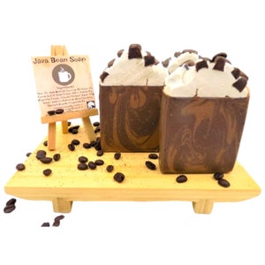 May include: Three bars of Java Bean Soap on a wooden tray with coffee beans scattered around. The soap is brown and white with a swirl pattern and coffee beans on top. A small easel with a sign that reads "Java Bean Soap" is in the background.