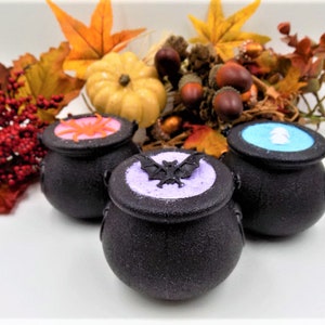 May include: Three black cauldron-shaped bath bombs with lids. The lids are decorated with Halloween-themed designs: a spider, a bat, and a ghost. The bath bombs are surrounded by fall foliage.
