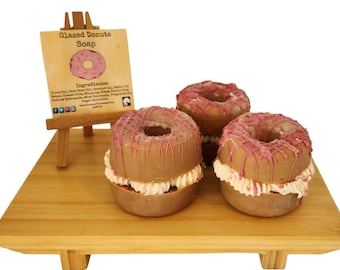 May include: Three donut-shaped soaps with pink frosting and white cream filling sit on a wooden surface. A small wooden easel displays a sign that reads "Glazed Donuts Soap" with a donut illustration.