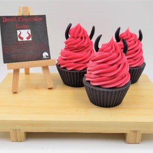 May include: Three devil cupcake soaps with red frosting and black horns sit on a wooden tray. A small chalkboard sign on a wooden easel displays the product name and ingredients. The cupcakes are in dark brown cupcake liners.